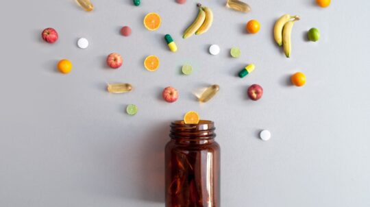 Vitamin pills and capsules alongside apples, bananas, lime and oranges coming out of a medicine jar, nutrition concept.