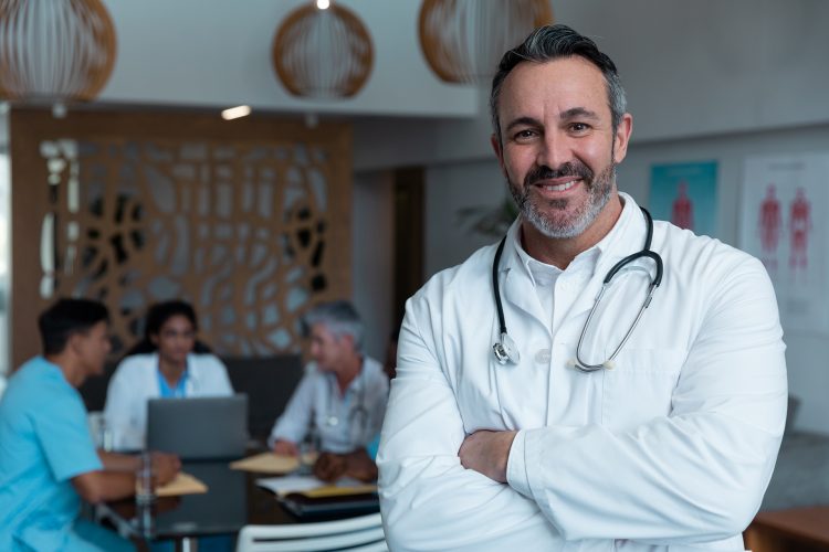 Portrait of smiling caucasian male doctor, with colleagues in discussion in the background. medicine, health and healthcare services.